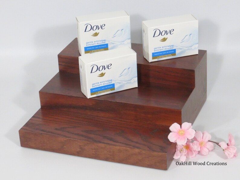 May include: Three Dove gentle exfoliating body wash bars displayed on a three-tiered wooden stand. The stand is made of dark wood with a reddish brown hue. The stand is on a white surface. The Dove bars are white with blue and gold accents. The text on the bars reads "Dove gentle exfoliating exfoliant douceur beauty bar - pain de beaut&eacute;"