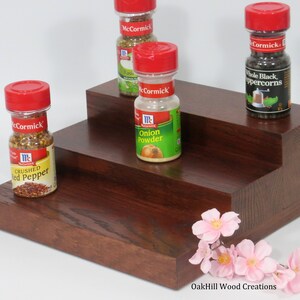 May include: A three-tiered wooden spice rack with four glass spice jars with red lids. The jars are labeled "McCormick" and contain various spices, including crushed red pepper, onion powder, basil, and whole black peppercorns. The rack is made of dark wood with a reddish brown stain. A sprig of pink flowers is in the bottom right corner of the image. The text "OakHill Wood Creations" is at the bottom of the image.