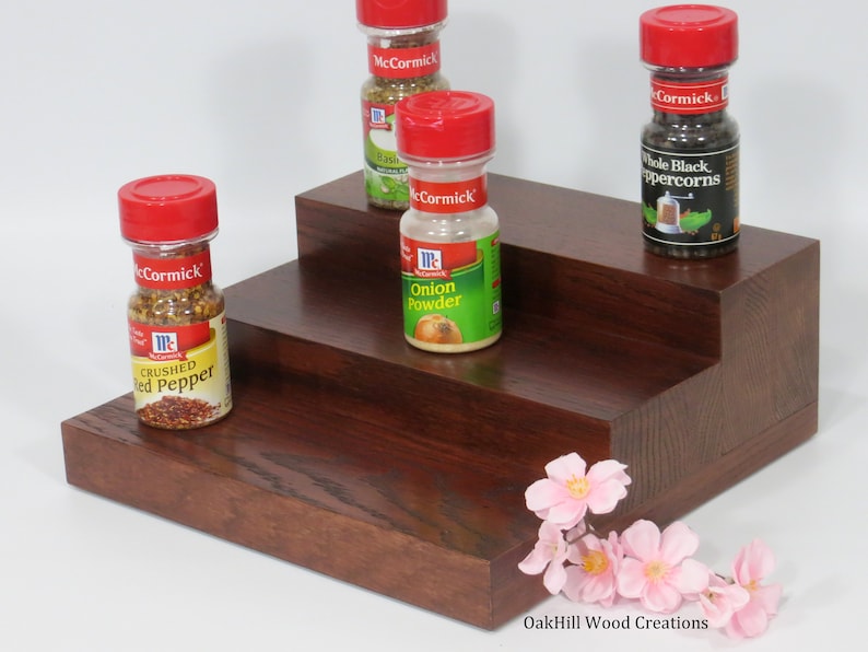 May include: A three-tiered wooden spice rack with four glass spice jars with red lids. The jars are labeled "McCormick" and contain various spices, including crushed red pepper, onion powder, basil, and whole black peppercorns. The rack is made of dark wood with a reddish brown stain. A sprig of pink flowers is in the bottom right corner of the image. The text "OakHill Wood Creations" is at the bottom of the image.