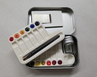 Perfect Pocket Paint Kit Watercolor Tin with 2 ceramic pallets, 18 wells for color, glass water jar, paintbrush and paper clip with magnets.