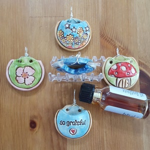 May include: A collection of handmade ceramic pendants with floral and whimsical designs. The pendants feature a variety of colors, including blue, green, pink, and red, and are suspended by silver wire. A small glass bottle with a dark liquid is also present.
