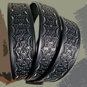 May include: A black leather belt featuring a repeating embossed pattern of skulls arranged within a geometric grid. The surface of the belt displays a detailed, textured design with deep impressions that highlight the skull motifs across the strap.