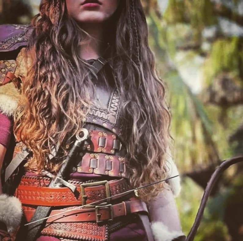 May include: A close-up of a person wearing a burgundy and brown leather breastplate with a sword and bow. The breastplate has intricate designs and is secured with belts and buckles. The person has long, wavy brown hair.
