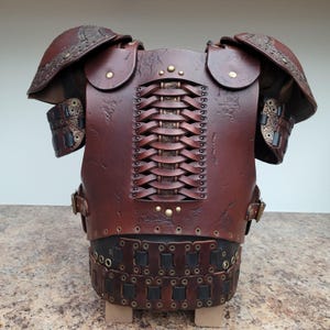 May include: A handcrafted, brown leather breastplate with intricate detailing. The armor features shoulder pauldrons, woven back panel, and decorative metal rivets. The design suggests a medieval or fantasy theme.