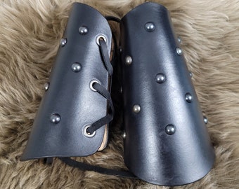 May include: A pair of black leather arm guards with silver-colored studs. The arm guards have a laced closure and are displayed on a brown, furry surface. The leather appears smooth and the studs are evenly spaced.