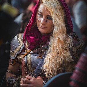 May include: A person wearing a burgundy hood and ornate armor with gold accents. The armor has a leather appearance with intricate detailing. The person has blonde hair and dark markings on their face, suggesting a costume or historical reenactment.