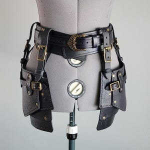 Puede incluir: Cinturón utilitario de cuero negro con hebillas de latón y detalles decorativos. El cinturón presenta dos bolsas y una hebilla central con un diseño ornamentado. El cinturón se muestra en un maniquí.