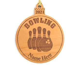 May include: A round, wooden Christmas ornament with a bowling theme. The ornament features the word "Bowling" above an illustration of bowling pins and a ball. The year "2021" is at the top, and "Name Here" is at the bottom. A gold string is attached for hanging.