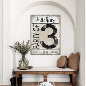 May include: A rustic wooden sign with the text "Hutchins Party of 3" in black lettering. The names Justin, Erin, and Alexis are listed below the number 3. The sign is hanging on a white wall in a hallway with a wooden bench and a vase of flowers.