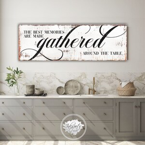 May include: White wooden sign with black text that reads "The best memories are made gathered around the table." The sign has a distressed, rustic look and is hanging on a wall above a kitchen counter.