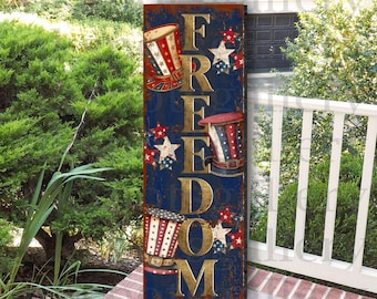 以下が含まれることがあります： 青い背景に金色の文字で「FREEDOM」と書かれた縦型の木製看板。看板は赤、白、青の星と、赤、白、青の帽子3つで飾られています。