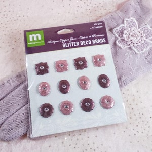 May include: A package of Glitter Deco Brads in various shapes and shades of pink and brown, with a glittery finish. The package is resting on a lavender lace glove with a decorative flower.