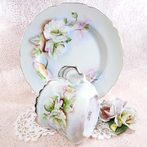 May include: A vintage teacup and saucer set with a delicate floral design. The cup and saucer are white with a pale blue background and feature hand-painted pink and white flowers with green leaves.