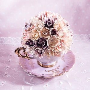 May include: A delicate bouquet of seashells arranged in a vintage teacup and saucer. The shells are in shades of white, pink, and purple, creating a soft and romantic look. The teacup and saucer are decorated with gold trim.