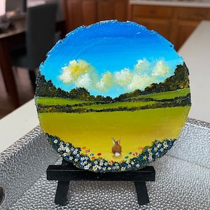 May include: A round wooden painting of a field with a blue sky, white clouds, green grass, and a small rabbit in the foreground. The painting is on a small easel.