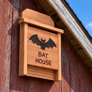 May include: A rectangular wooden bat house with a natural light brown finish. The front panel features a black silhouette of a bat with wings spread and the text BAT HOUSE engraved in dark lettering. The structure has a slatted base.