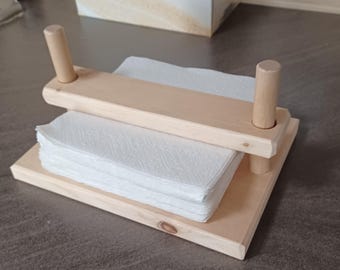 May include: A light-colored wooden napkin holder with a rectangular base and a top piece held up by two wooden dowels. The holder contains a stack of white paper napkins. The design is simple and functional.