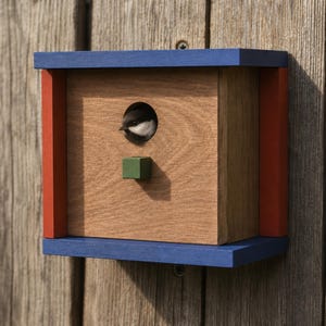 Könnte beinhalten: Ein Vogelhaus aus Holz mit blauem Ober- und Unterteil, roten Seitenakzenten und einer Vorderseite aus Naturholz. Ein kleiner Vogel schaut aus dem kreisförmigen Eingang. Unter dem Eingang befindet sich ein kleiner grüner Würfel. Das Vogelhaus ist an einer Holzwand befestigt.