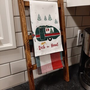 May include: A wooden ladder style towel rack with a white and red checked towel and a white towel with a green camper and the text "Deck the Haul" hanging on it.