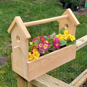 May include: A wooden planter box with a handle, shaped like a birdhouse, with pink and yellow flowers planted inside.