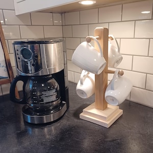 May include: A wooden mug rack with four white ceramic mugs hanging from it. The rack is made of natural wood and has a simple design. The rack is sitting on a black countertop next to a silver coffee maker.