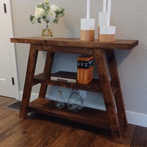 Puede incluir: Una mesa consola de madera marrón oscuro con dos estantes. La mesa tiene un estilo rústico, de granja y presenta un acabado desgastado. La mesa está decorada con un jarrón de flores blancas, dos candelabros blancos y una pila de libros.