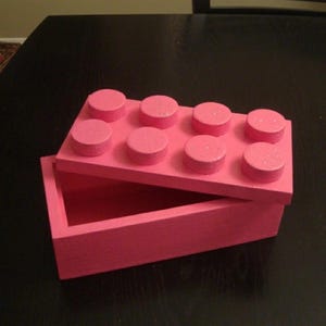 May include: A pink wooden box designed to resemble a large building block. The lid features eight raised circular studs, and the box is open, revealing an empty interior. The box is painted in a solid pink color.