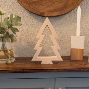 May include: A wooden Christmas tree ornament with a layered design. The tree is made of light-coloured wood and stands on a dark wooden surface. A white candle and a vase with flowers are also visible.
