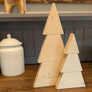 May include: Two wooden Christmas tree decorations. The trees are made of light-colored wood and are assembled from triangular and rectangular pieces. The trees are displayed on a wooden surface, with a white ceramic jar in the background.