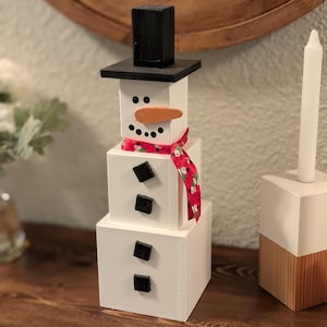 May include: A wooden snowman decoration with a black top hat, a carrot nose, and a red scarf with a white and green pattern. The snowman has black square buttons and a candle holder is next to it.