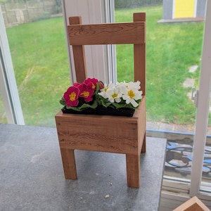 May include: A wooden chair-shaped planter holding pink and white flowers. The planter is constructed from light brown wood, showcasing a rustic aesthetic. The flowers are in full bloom, providing a splash of colour to the setting.