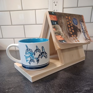 May include: A wooden book stand with a book titled "Water for Elephants" by Sara Gruen. A white ceramic mug with a blue interior and a design of blue and black orcas is on the stand.