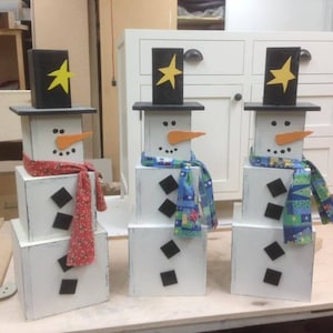 以下が含まれることがあります： 白い塗料で塗られた箱を積み重ねて作った、木製の雪だるまの飾り3つ。雪だるまは、黄色い星がついた黒い帽子をかぶり、オレンジ色の鼻と黒い目をしています。カラフルなマフラーをしています。