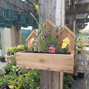 Op de afbeelding: Een houten plantenbak met een huisvormig ontwerp, met verschillende bloemen, waaronder gele narcissen, roze anjers en paars-gele viooltjes. De plantenbak is bevestigd aan een houten paal.