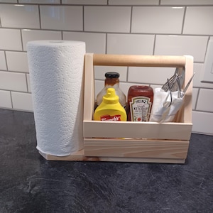 Pode incluir: Um caddy de madeira com uma alça, um suporte para papel toalha e um espaço para condimentos. O caddy contém duas garrafas de vidro de condimentos, uma amarela e uma vermelha, e um copo transparente com talheres. O caddy é feito de madeira natural e tem um visual rústico.