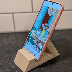 May include: A smartphone in a coral case rests on a wooden phone stand. The phone screen shows a digital illustration of a dining room, featuring a table set with tableware and a vase of flowers. The stand is made of natural wood.