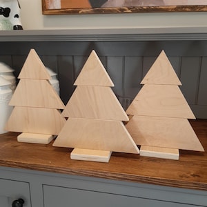Peut inclure: Trois sapins de Noël en bois, chacun composé de trois pièces de bois triangulaires empilées, sont exposés sur une surface en bois. Les arbres sont tous de la même taille et de la même forme, et ils sont tous en bois clair.