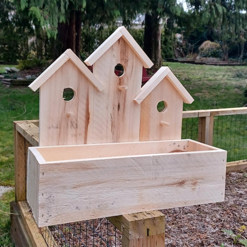 Op de afbeelding: Een houten vogelhuisje-plantenbak met drie vogelhuisjes bovenop en een rechthoekige plantenbak eronder. De vogelhuisjes zijn gemaakt van natuurlijk hout en hebben kleine openingen waar vogels naar binnen kunnen. De plantenbak is ook gemaakt van natuurlijk hout en heeft een lichtbruine afwerking.