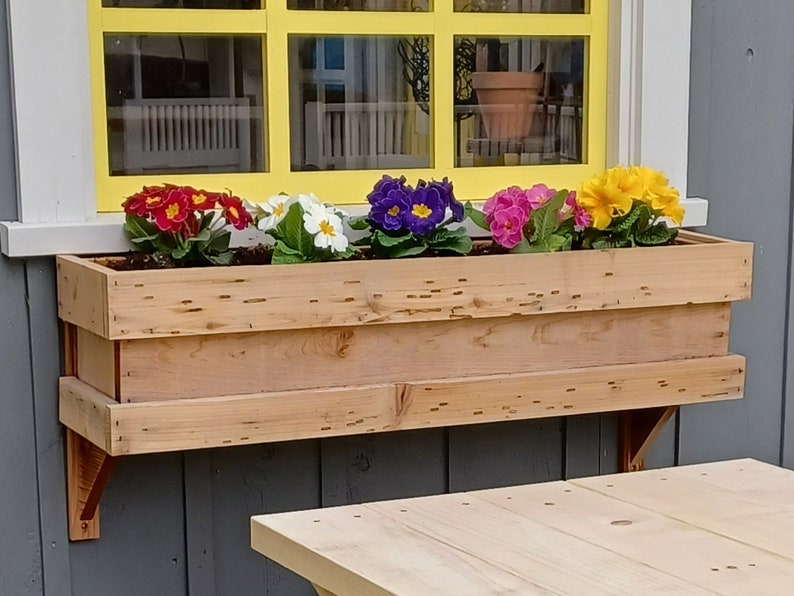 May include: A wooden window box planter with a variety of colorful flowers, including red, white, yellow, purple, and pink. The planter is mounted on a gray wall with a wooden bracket.