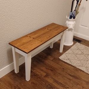 May include: A white wooden bench with a stained wood top. The bench has a simple design with straight legs and a rectangular top. The bench is in a hallway with a white tiled floor and a white umbrella stand.