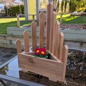 May include: A wooden planter with a triangular base and vertical picket-style accents. The planter is filled with vibrant red and yellow flowers. The wood has a natural finish, and the design is suitable for outdoor use.