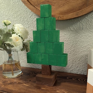 May include: A handcrafted wooden Christmas tree decoration. The tree is constructed from stacked green blocks, with a brown trunk and base. The tree is displayed on a wooden surface, with a vase of flowers in the background.
