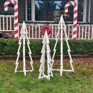 May include: Three white wooden Christmas tree frames, each with a different size and shape, standing on a green lawn. The trees are decorated with red and white candy canes and a red and white wreath.