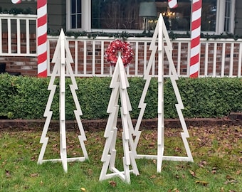 Puede incluir: Tres marcos de árbol de Navidad de madera blanca, cada uno con un tamaño y una forma diferentes, de pie en un césped verde. Los árboles están decorados con bastones de caramelo rojos y blancos y una corona roja y blanca.