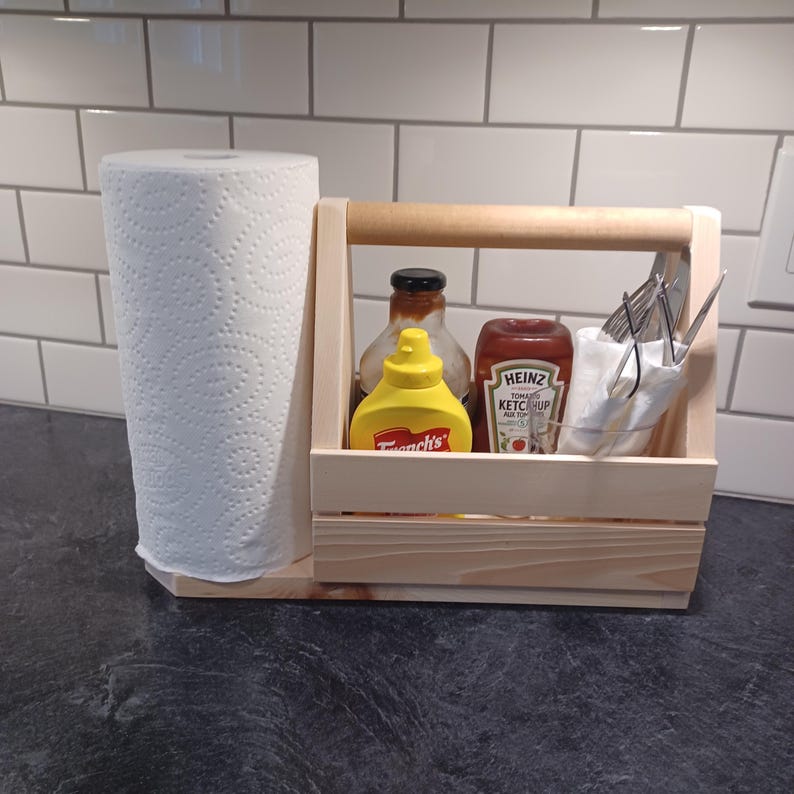 K&ouml;nnte beinhalten: Ein Holz-Caddy mit Griff, einem Papierhandtuchhalter und Platz f&uuml;r Gew&uuml;rze. Der Caddy enth&auml;lt zwei Glasflaschen mit Gew&uuml;rzen, eine gelbe und eine rote, sowie ein klares Glas mit Besteck. Die Flaschen tragen die Etiketten "French's" und "Heinz Tomato Ketchup".