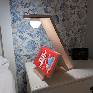 May include: A wooden desk lamp with a built-in book holder. The lamp has a warm, natural wood finish and a bright, round light. A red book titled "The Retirement Plan" is displayed in the holder. A digital clock displays the time as 4:09.