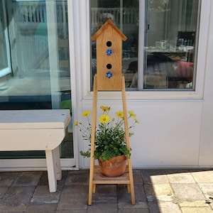 Könnte beinhalten: Ein hölzernes Vogelhaus mit einem dekorativen blauen Blumendesign, montiert auf einem hölzernen Pflanzenständer. Der Ständer hält einen Terrakottatopf mit grünen Pflanzen und gelben Blumen. Das Vogelhaus hat drei Öffnungen.