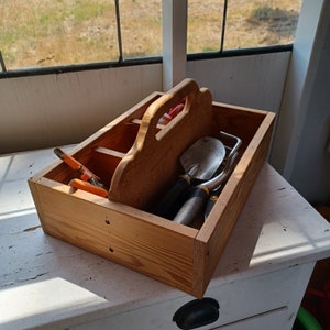 May include: A wooden gardening tool caddy with a handle. The caddy is filled with gardening tools, including a trowel, a hand rake, and a pair of pruning shears.