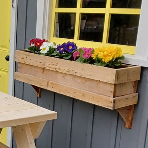 May include: A wooden window box planter with a variety of colorful flowers, including red, white, pink, purple, and yellow. The planter is mounted on the exterior wall of a building.
