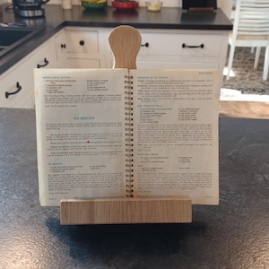 May include: A wooden cookbook stand holds an open cookbook on a kitchen countertop. The cookbook is open to a recipe for "Tea Biscuits." The stand is made of light-colored wood and has a simple, functional design.